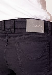 Pantalones de mezclilla oscura con un corte ajustado, con una etiqueta de cuero gris marcada con "Jeckerson" y costuras inclinadas en los bolsillos traseros.