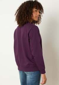 Sudadera morada de tejido suave, con un cuello redondo y puños de canalé. Llevada con vaqueros de color azul claro, vista desde atrás.