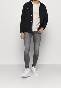 Chaqueta negra de botones sobre una camiseta de color beige claro, acompañada de jeans ajustados grises y zapatillas blancas, con un diseño elegante y estilo casual.