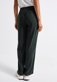 Mörkgröna vida byxor med en samlad elastisk midja, sidofickor och en mjuk tygstruktur. Bärs med vita sneakers.