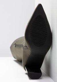 Botín de tobillo verde oliva con punta afilada, suela de goma negra texturizada y detalle de cremallera lateral. Material suave con un acabado mate.