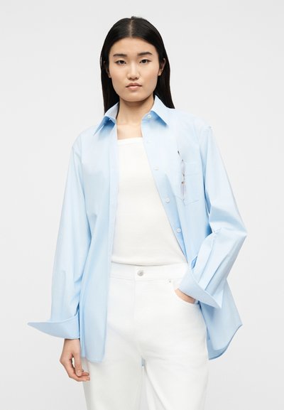 Γυναίκα με ίσια σκουρόχρωμα μαλλιά φορά ανοιχτό γαλάζιο πουκάμισο πάνω από λευκή μπλούζα και λευκό παντελόνι, με γυαλιά που είναι στερεωμένα στην τσέπη του πουκαμίσου.