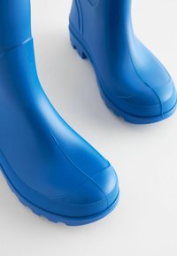 Kék gumicsizmák sima felülettel, lekerekített orral, texturált talppal és ívelt felső résszel. Enyhén fénylő és modern dizájn jellemzi őket.