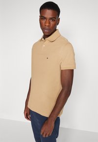 Beige polo ing klasszikus gallérral, rövid ujjal és apró logóval a mellkason. Sötétkék nadrággal viselve.