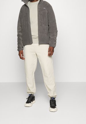 Mand iført grå fleecejakke med lynlås, lysebeige bukser og sorte og hvide sneakers, stående mod en ensfarvet lys baggrund.