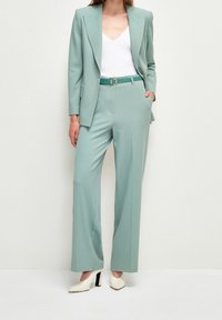 Jasnozielony garnitur z dopasowaną marynarką i szerokimi spodniami, biała bluzka z dekoltem w serek oraz białe buty na blokowym obcasie. Zielony pasek z metalową klamrą.