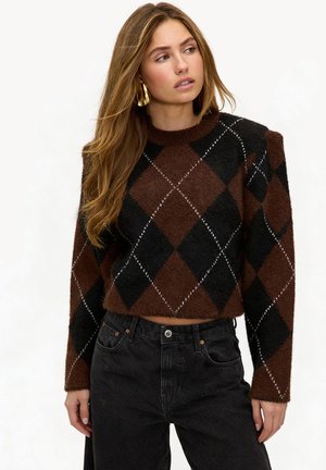 Vrouw met lang haar die gouden creolen draagt, een bruin-zwart argyletrui en zwarte jeans met hoge taille, kijkt opzij.