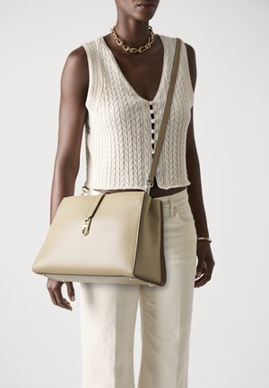Femme portant un haut sans manches en tricot torsadé crème et un pantalon, collier chaîne dorée, sac bandoulière beige avec fermoir doré.