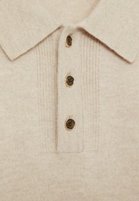 Polo en maille beige avec un col, présentant trois boutons ronds marron et des détails texturés subtils sur le plastron.
