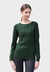 Pull en maille torsadée vert avec des manches longues, un col rond et un ourlet côtelé. Fabriqué dans un tissu doux et texturé. Porté sur un jean noir.
