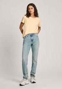 Dzeltena īsām piedurknēm T-kreklā ar apaļu kakla izgriezumu, savienots ar gaiši ziliem augstās jostas vietas džinsiem un baltām sporta kurpēm. Tekstūrēta auduma, ikdienišķa piegriezuma.