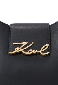 Black leather handbag featuring a gold-tone cursive "Karl" logo on the flap, with smooth texture and clean edges.