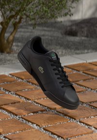 Baskets en cuir noir avec un design à lacets, présentant un petit logo vert et un marquage sur le côté. Semelle texturée sur une surface en bois.