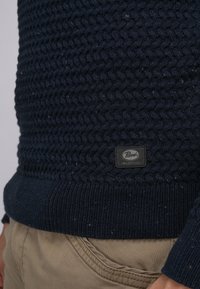 Pulover tricotat bleumarin cu un model texturat țesut, tiv ondulat și etichetă din material negru cu inscripția "Parol Industries". Pantaloni bej parțial vizibili.