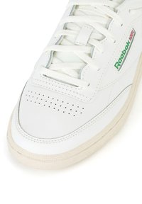 Balti odiniai sportbačiai su perforuota nosimi, plokščiomis raišteliais ir žalia logo detale. Paminkštintas padas su rusvu gumuotu tekstūra ir siuvinėjimo akcentais.