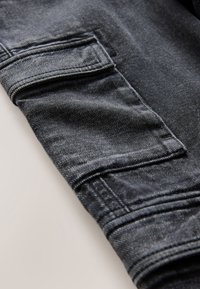 Primo piano di tessuto denim grigio scuro con una tasca rettangolare con patta e cuciture doppie visibili sulla manica.