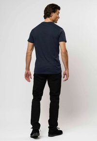 Férfi áll, háttal felénk, sötétkék, rövid ujjú pólót, fekete nadrágot és fekete cipőt visel, egy sima, világos háttér előtt.