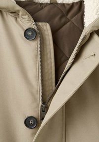 Abrigo beige con cremallera frontal y cierre de botón. El interior cuenta con un forro acolchado marrón y una tela suave y texturizada.