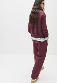 Felpa in pile color bordeaux con vestibilità rilassata, indossata sopra una camicia a righe. Abbinata a pantaloni coordinati e scarpe slip-on. Texture morbida, design casual.