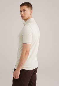 Short-sleeve polo shirt in light beige, textured fabric, with a classic collar and a slim fit design. Worn with dark brown pants.