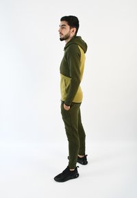 Chándal verde oliva con capucha, que presenta un diseño de bloques de color y bolsillos laterales, combinado con zapatillas deportivas negras. Textura suave.