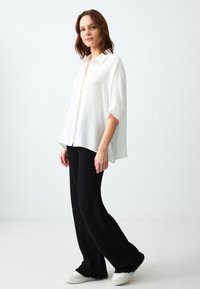 Chemise blanche à manches courtes et fluides, associée à un pantalon large texturé noir et des baskets blanches sur un fond neutre.