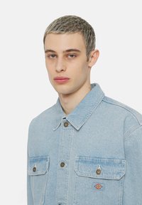 Ljusblå denimjacka med spetsig krage, två bröstfickor med knappstängning och en liten logo på vänster ficka.