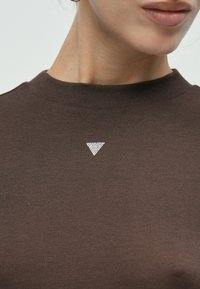 Καφέ πλεκτό τοπ με ριμπ ύφασμα, διακοσμημένο με τριγωνικό στρας στο λαιμό. Μανίκια κοντά και σχέδιο με ψηλό γιακά.