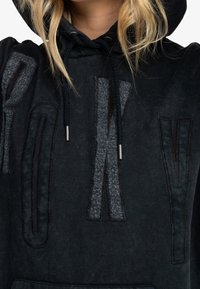 Hoodie noir avec de grandes lettres texturées. Fabriqué dans un matériau en feutre doux avec une finition grise contrastante. Comprend des cordons et des poches.