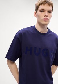 Tamsiai mėlynas medvilninis marškinėlis trumpomis rankovėmis ir apvaliu kaklu. Krūtinės srityje yra iškilus "HUGO" logotipas, kuris yra tamsesnio atspalvio mėlynos spalvos.