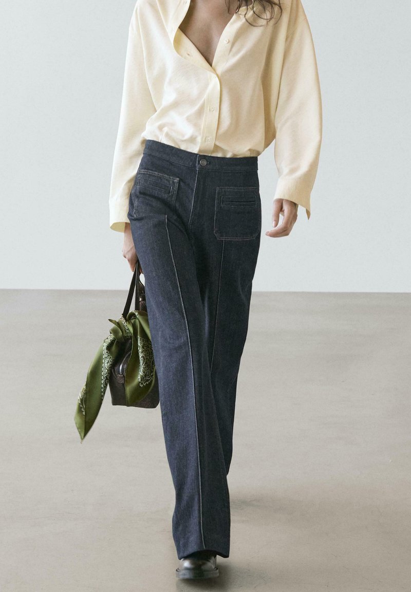 Dunkelblaue Jeans im weiten Bein mit kontrastierenden Nähten, kombiniert mit einem hellgelben Hemd und einer braunen Tasche mit grünem Schal.