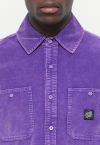 Purple corduroy shirt featuring a classic collar, buttoned front, two chest pockets, and a small logo patch on the right pocket.