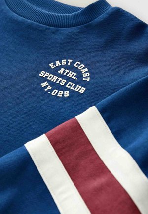 Modrá mikina s úpletem kolem krku, se bílým nápisem "EAST COAST ATHL. SPORTS CLUB NY.025" a bílou-červenou-bílou pruhovanou látkou na rukávu.