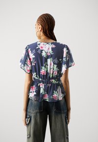 Blomstrete peplum topp i marineblå med samlet midje. Har korte, flagrende ermer og et gjennomsiktig stoff med rosa og grønne blomster-mønstre.