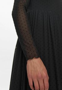 Rochie neagră cu mâneci lungi dinmaterial transparent cu buline, țesătură texturată și fustă fluidă, având un tiv ondulat la marginea mânecilor.
