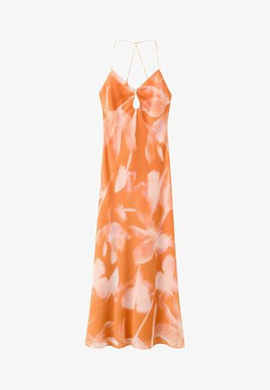 Femme aux cheveux bouclés portant une robe dos nu orange et rose à fines bretelles et des sandales compensées noires, debout sur fond blanc.