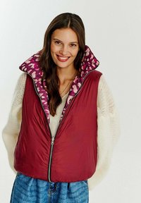 Červená vycpaná vesta s vysokým límcem a kontrastní purpurovou květinovou podšívkou. Přední zapínání na zip a uvolněný střih. Svetřík pod tím.