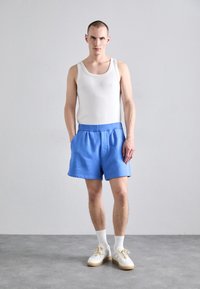 Top sin mangas blanco, pantalones cortos azules con bolsillos laterales y zapatillas blancas con suela beige. La tela tiene una textura suave y un corte relajado.