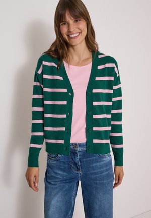 Smilende kvinde iført en grøn og lyserød stribet knappet cardigan over en lyserød top med blå jeans, stående foran en ensfarvet baggrund.