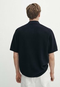 Man met kort lichtbruin haar in een zwart gestructureerd shirt met korte mouwen en witte broek, met de rug naar de camera tegen een effen witte achtergrond.