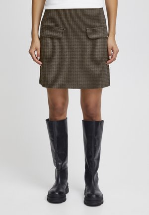 Kobieta w brązowej mini spódnicy z fakturą, z przednimi kieszeniami i czarnych wysokich kozakach, stojąca na tle jednolitego tła.