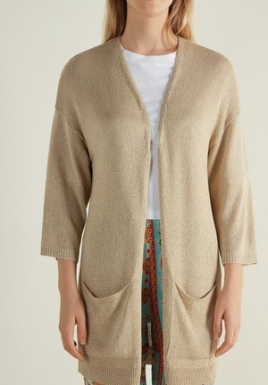Femme portant un cardigan beige en maille ouvert sur le devant avec poches, un t-shirt blanc et un pantalon bleu-rouge à motifs, debout sur un fond uni.