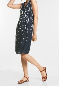 Marinblå långklänning utan ärmar med vitt fjärils mönster, knälång, i kombination med bruna sandaler med remmar. Slätt tyg, avslappnad passform.