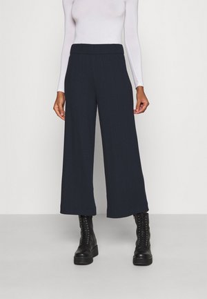 Kvinde iført marineblå vidde-cropped bukser, hvid langærmet top og sorte snørestøvler med plateau, stående på en ensfarvet baggrund.