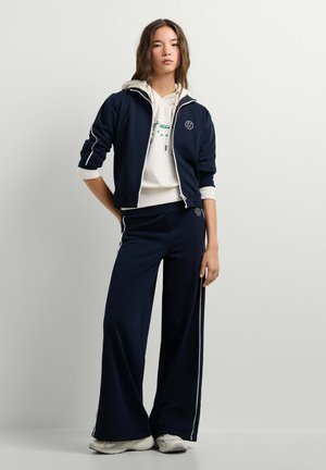 Vrouw staand, gekleed in een marineblauw trainingsjack en broek met witte strepen, een crème hoodie en witte sneakers tegen een effen achtergrond.