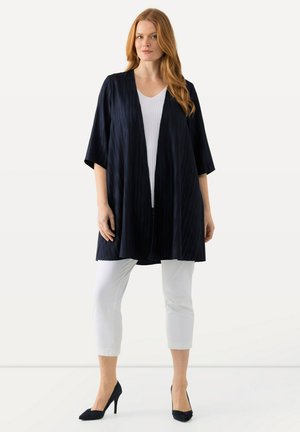 Kimono blu navy aperto davanti con un motivo testurizzato, abbinato a pantaloni bianchi a vita alta e tacchi neri. Il tessuto cade in modo morbido.