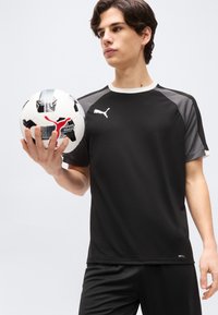 Νεαρός άνδρας με μαύρα αθλητικά ρούχα Puma κρατά μια λευκή και μαύρη μπάλα ποδοσφαίρου με κόκκινο λογότυπο Puma, σε απλό φόντο.