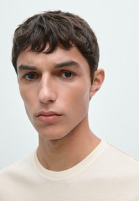 Lys beige t-shirt med rund hals, glat tekstur, tætsiddende design, subtile syningsdetaljer ved skulderen, ensfarvet baggrund.