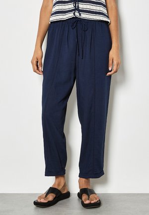 Kobieta w granatowych spodniach ze ściągaczem, podwiniętymi nogawkami i czarnych sandałach z odkrytymi palcami, stojąca na tle białej ściany.