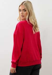Felpa rossa con una vestibilità ampia, maniche lunghe, polsini e orlo a coste. Texture liscia, design minimalista, silhoutte oversize.